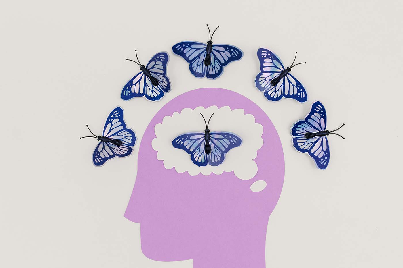 Illustration of a purple human head silhouette with butterflies flying around and inside the brain area, symbolizing thoughts, mental health, creativity, or mindfulness.
