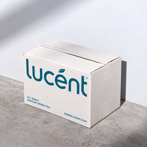 A white cardboard box of Lucent matcha green tea, featuring the brand name in teal blue text, set on a gray concrete floor against a white wall with soft shadows.