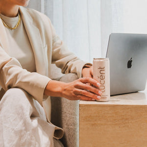 Person holding a can of Lucent energy drink in front of a laptop on a wooden table.