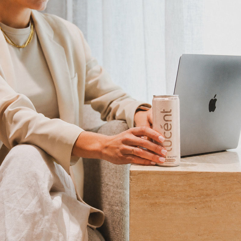 Person holding a can of Lucent energy drink in front of a laptop on a wooden table.